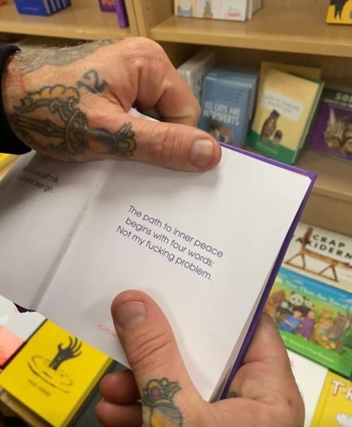 A person with tattooed hands holds open a small purple book in a bookstore, revealing a page with the text 'The path to…