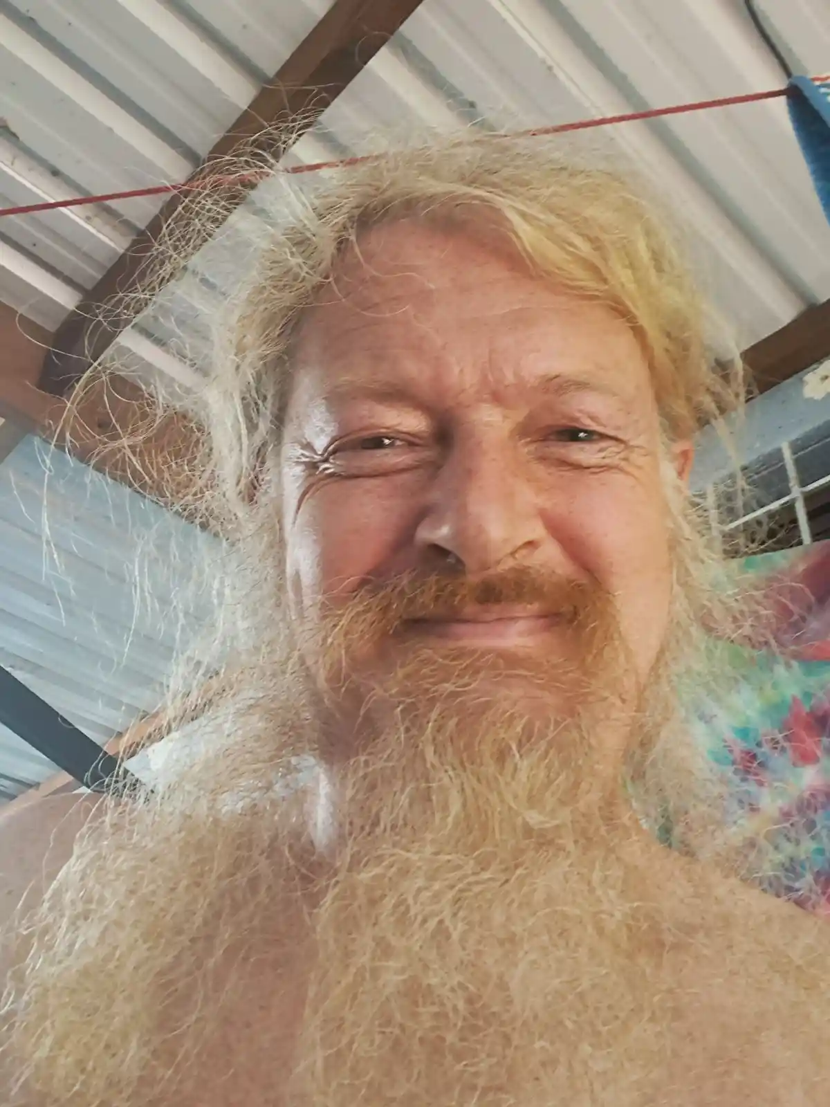 A close-up selfie of a man with long, shaggy blonde hair and a full, similarly colored beard, smiling slightly while looking…