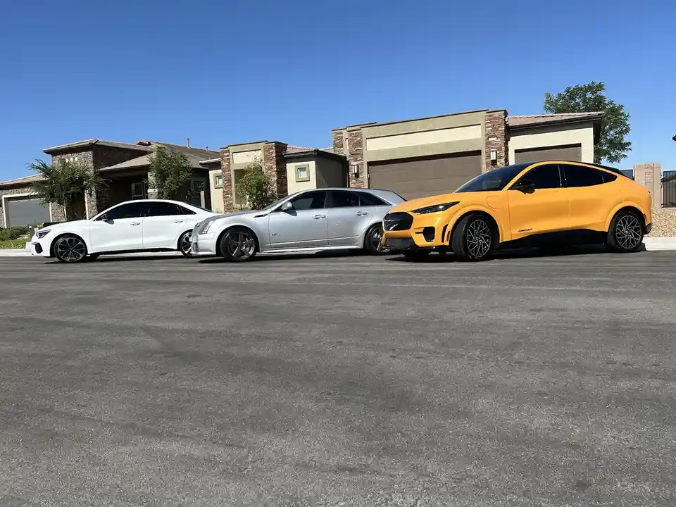 Three luxury cars—a white sedan, a silver station wagon, and a bright orange SUV—are parked side-by-side in a residential…