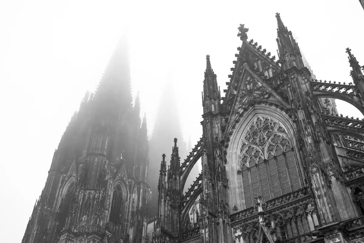 A black-and-white photograph of the Cologne Cathedral, its Gothic spires and intricate stonework partially obscured by mist…