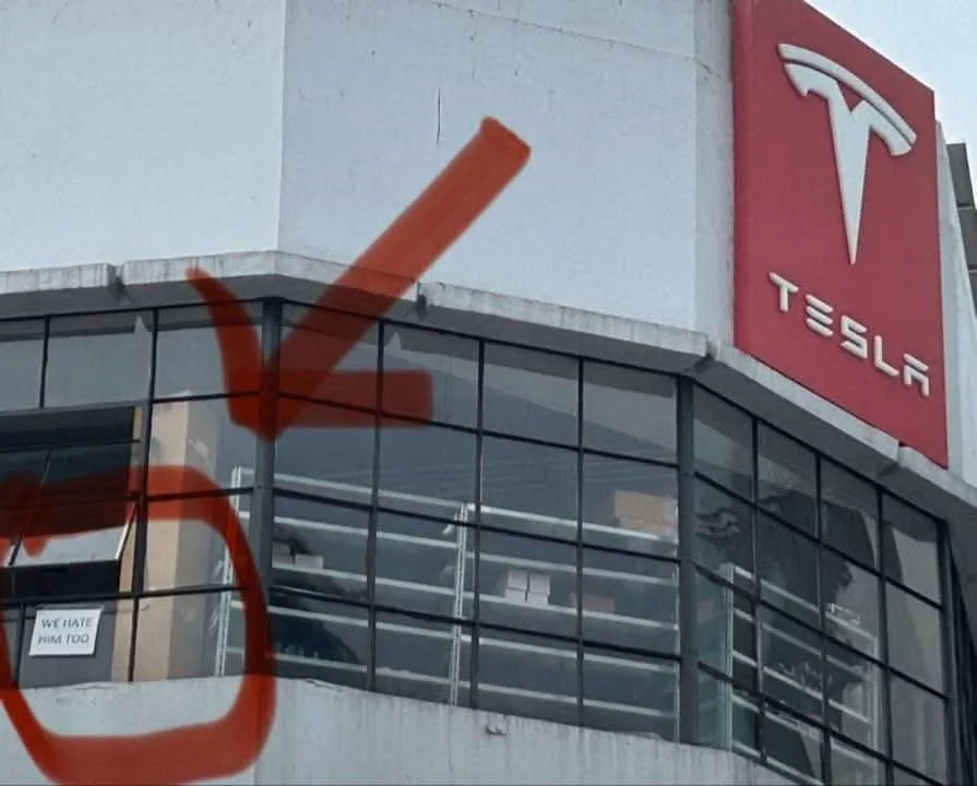 A photo of a Tesla building with a large red spray-painted arrow pointing to a small white sign on the building that reads…