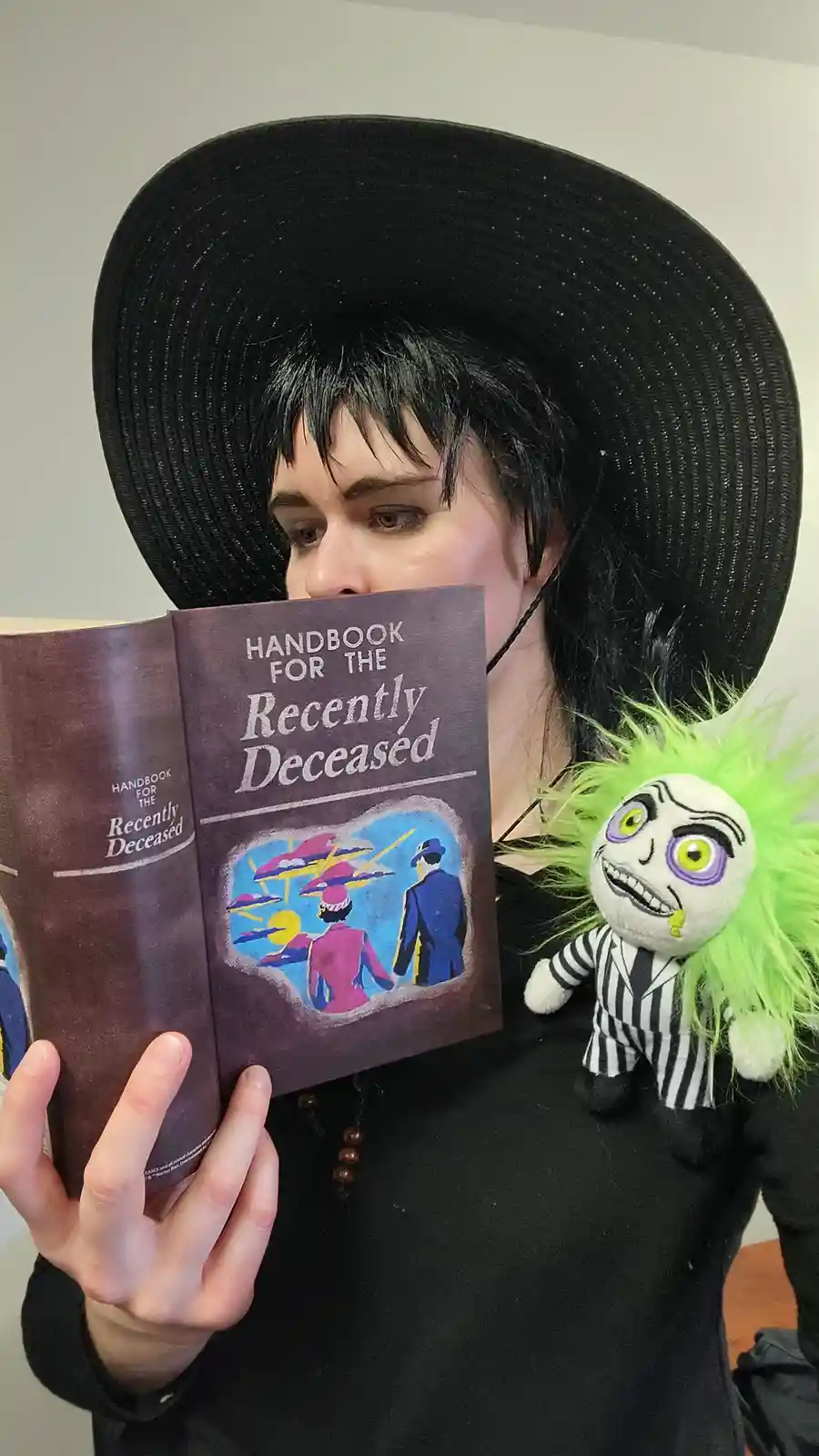 A person dressed as Wednesday Addams, wearing a large black hat and black clothing, holds up a book titled 'Handbook for the…