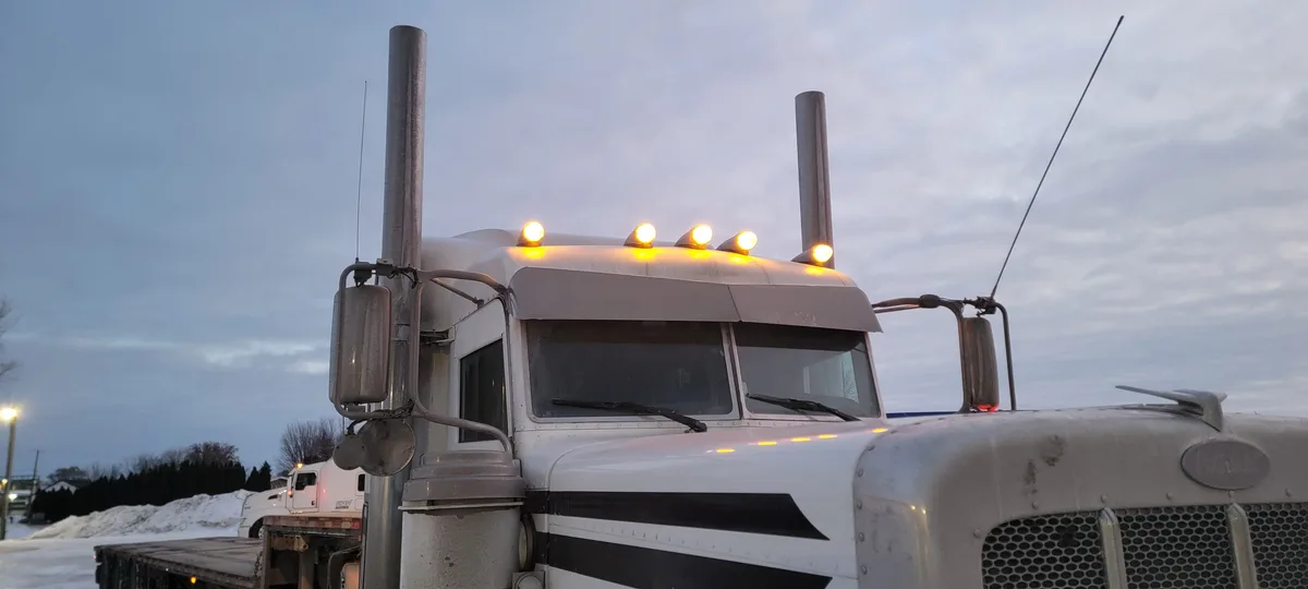 A white semi-truck with its roof lights on is parked in a snowy area during dusk or dawn. The truck has prominent chrome…