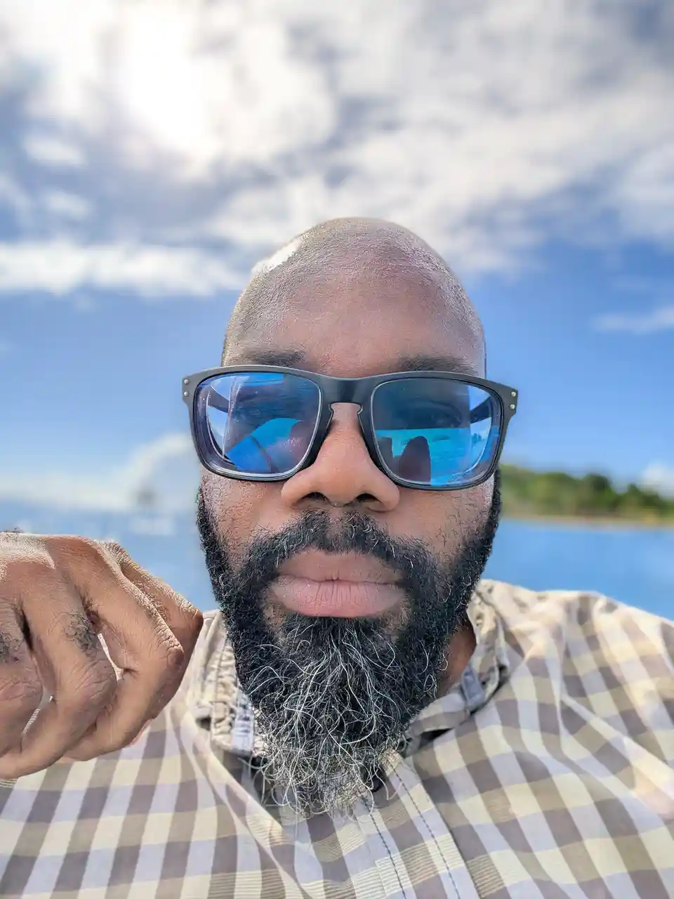 A close-up selfie of a bald man with a full beard showing gray hairs, wearing large black-framed sunglasses with blue…