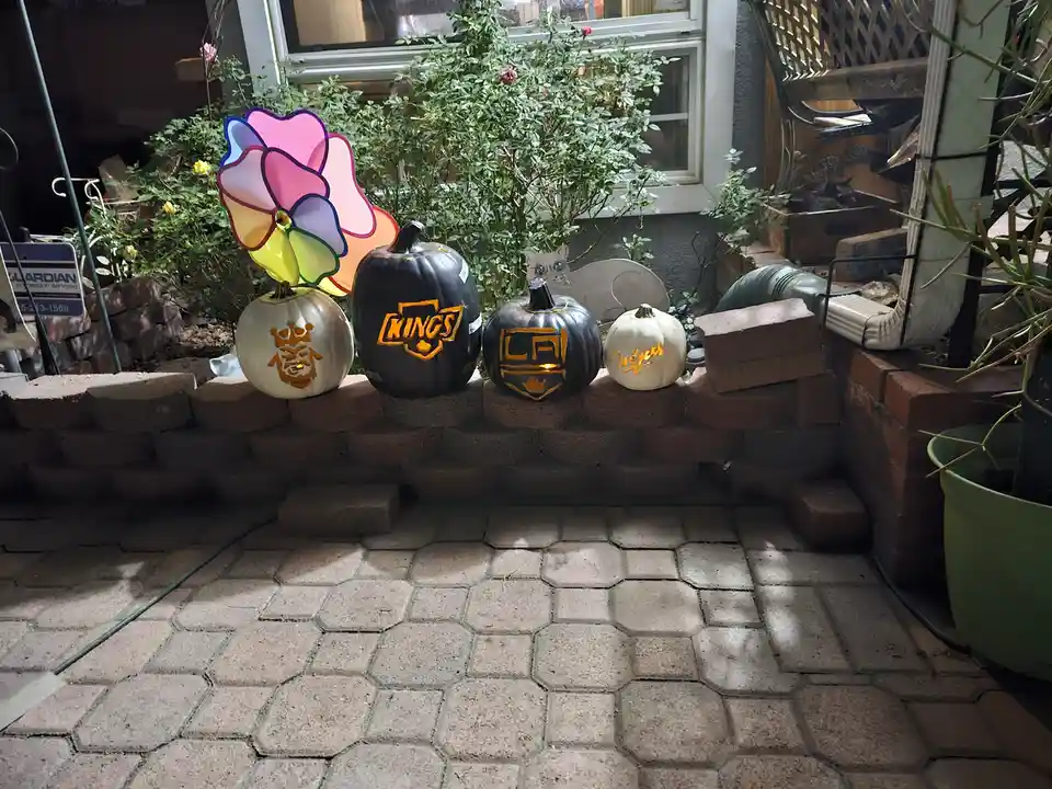 Four pumpkins sit on a brick ledge in a backyard, each decorated with a different Los Angeles sports team logo: one with a…