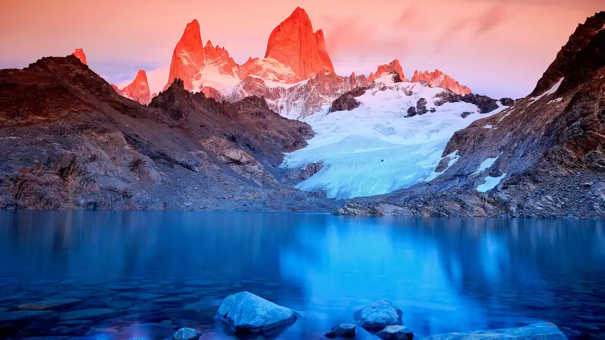 Monte Fitz Roy, a striking peak, glows with hot pink and deep blue hues as sunrise ignites the Argentine landscape. The…
