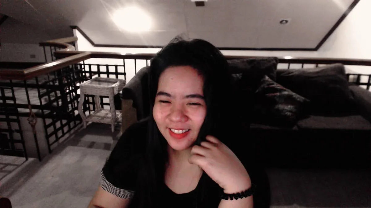 A young woman with dark hair, wearing a black shirt with striped sleeves, is smiling broadly while winking and touching her…