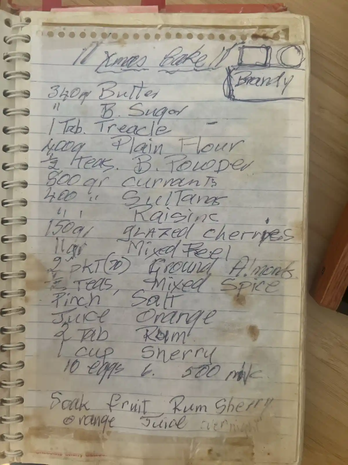 A handwritten recipe in a spiral-bound notebook titled 'Christmas Bake It' with a drawing of a bottle labeled 'Brandy' next…