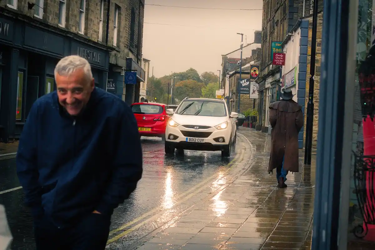 A man in a blue jacket smiles broadly while standing on a wet, rainy street with cars passing by. In the background, another…