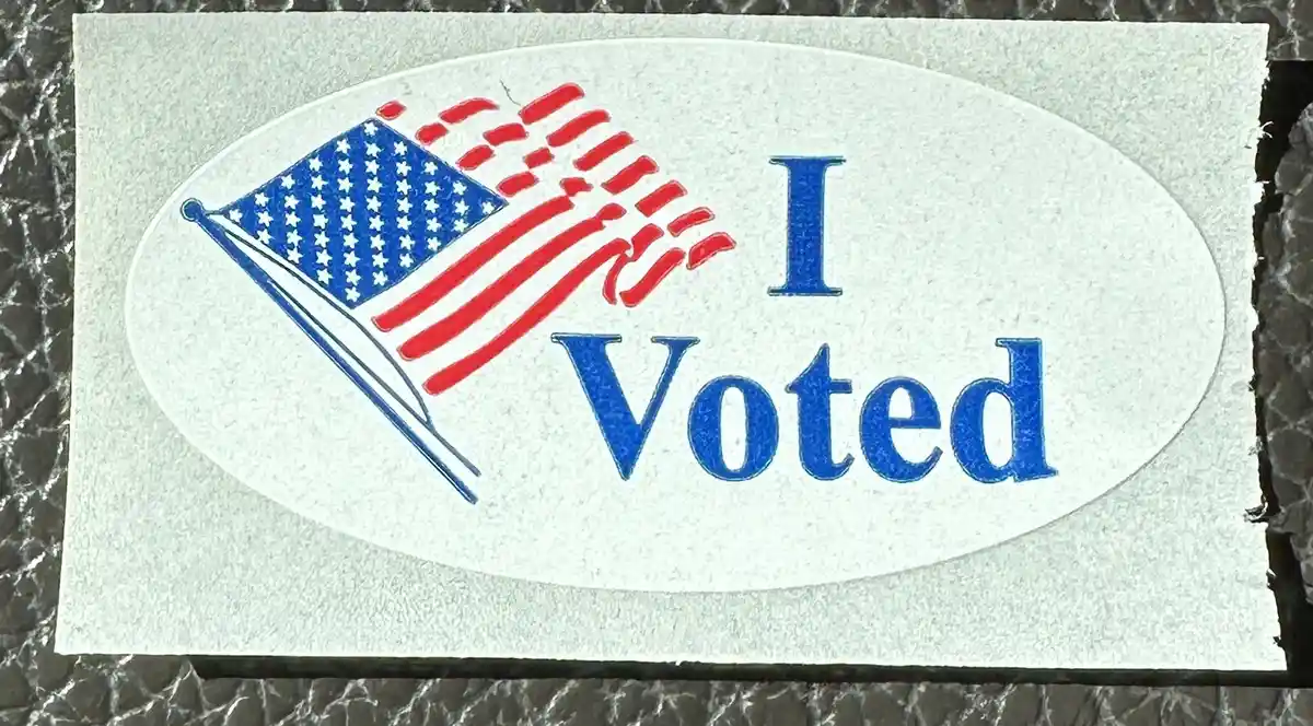 A close-up photo of a white oval sticker with a blue and red American flag illustration on the left and the text 'I Voted'…