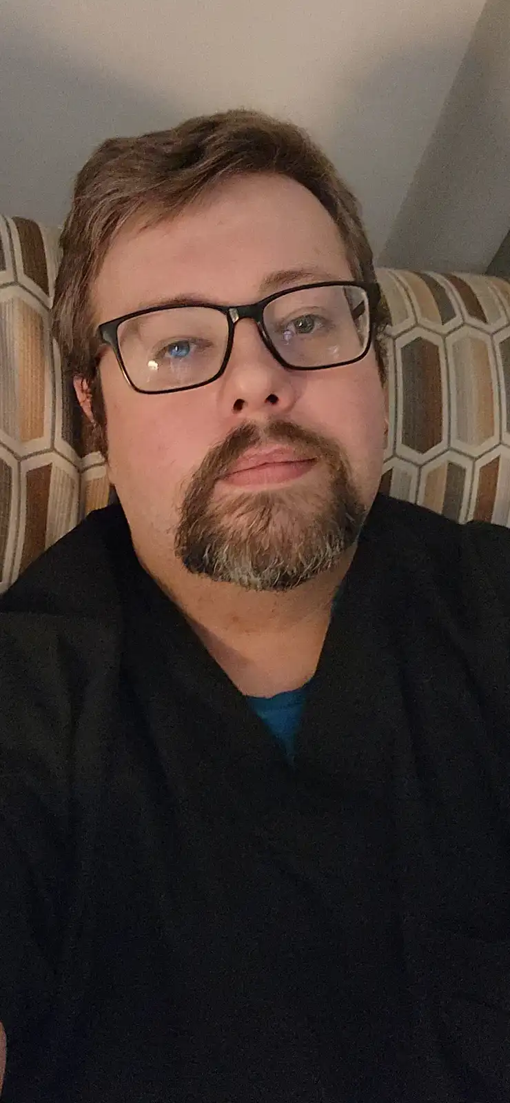 A man with glasses and a goatee sits on a patterned couch, looking directly at the camera with a neutral expression. The…