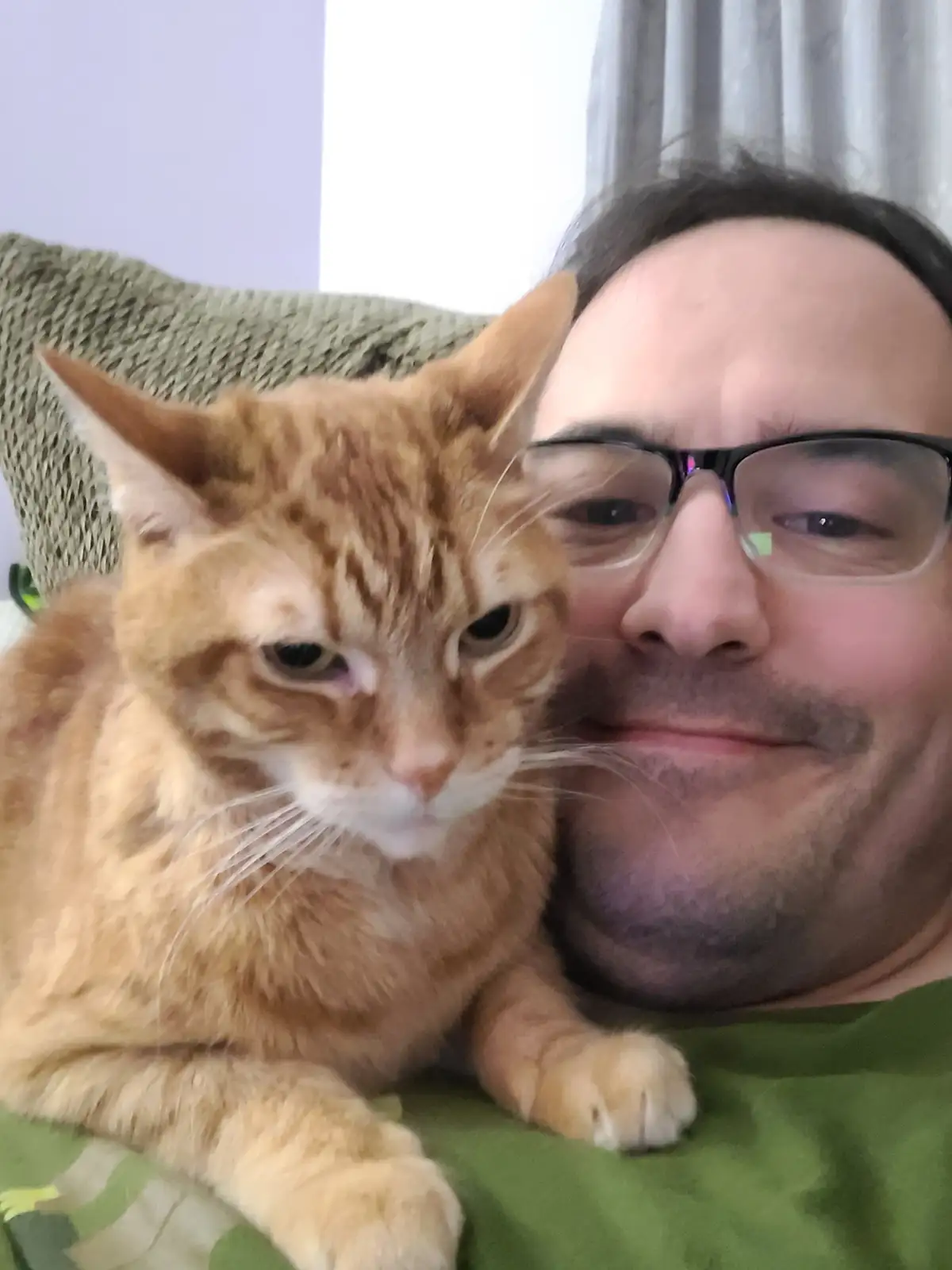 A selfie shows a man with glasses smiling while an orange tabby cat with a bored, unimpressed expression rests beside him…