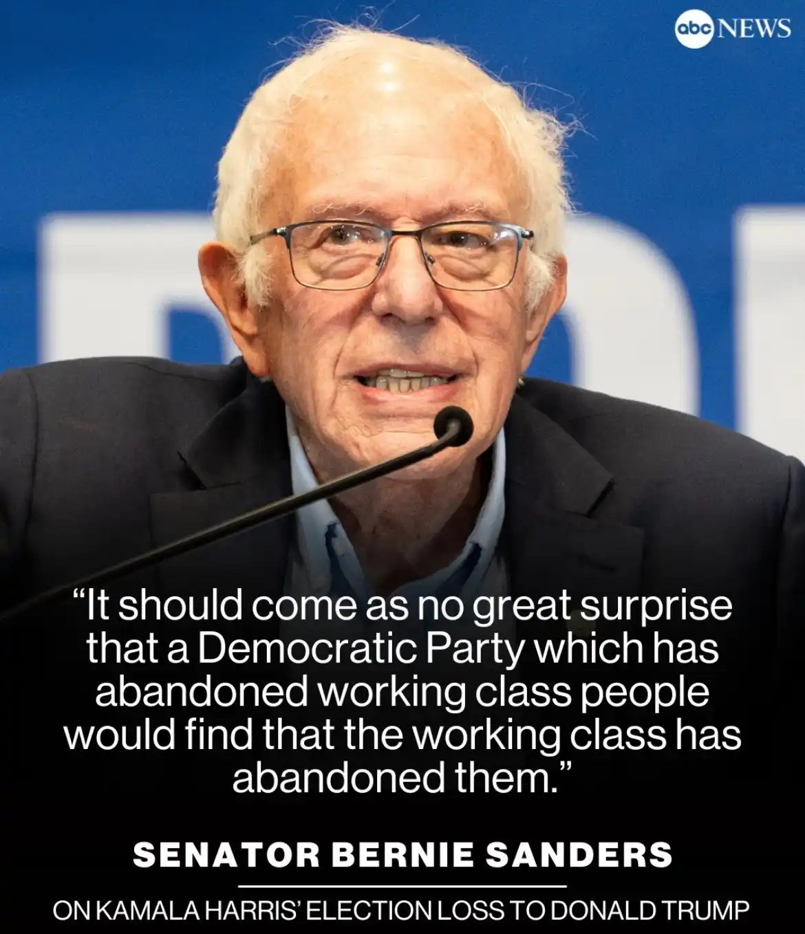 A close-up photo of Senator Bernie Sanders speaking at a press event, wearing glasses and a dark suit, with a microphone in…