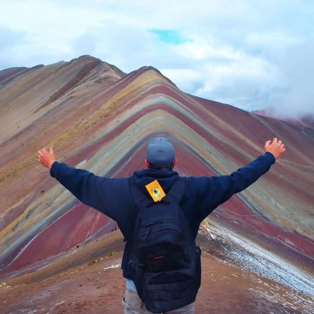 A person wearing a blue hoodie, grey cap, and black backpack stands with arms outstretched, facing a colorful, striped…