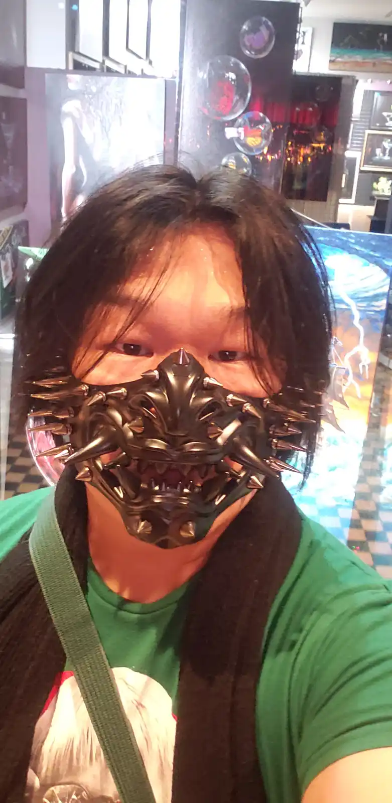 A person with long dark hair wears a black, spiked, dragon-like mask covering their mouth and nose, revealing only their…