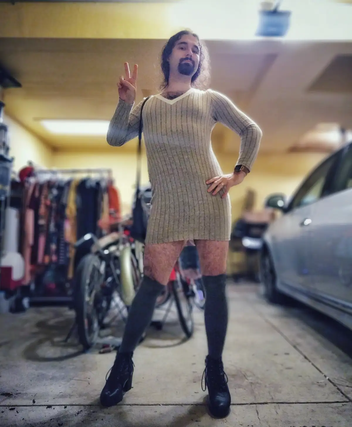 A man with long hair and a beard stands in a garage wearing a tight, ribbed, beige sweater dress that ends above his knees…