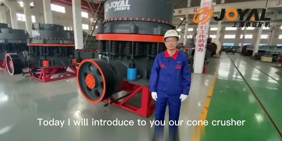 A man in a blue work uniform and white hard hat stands in a factory next to a large industrial cone crusher machine. The…