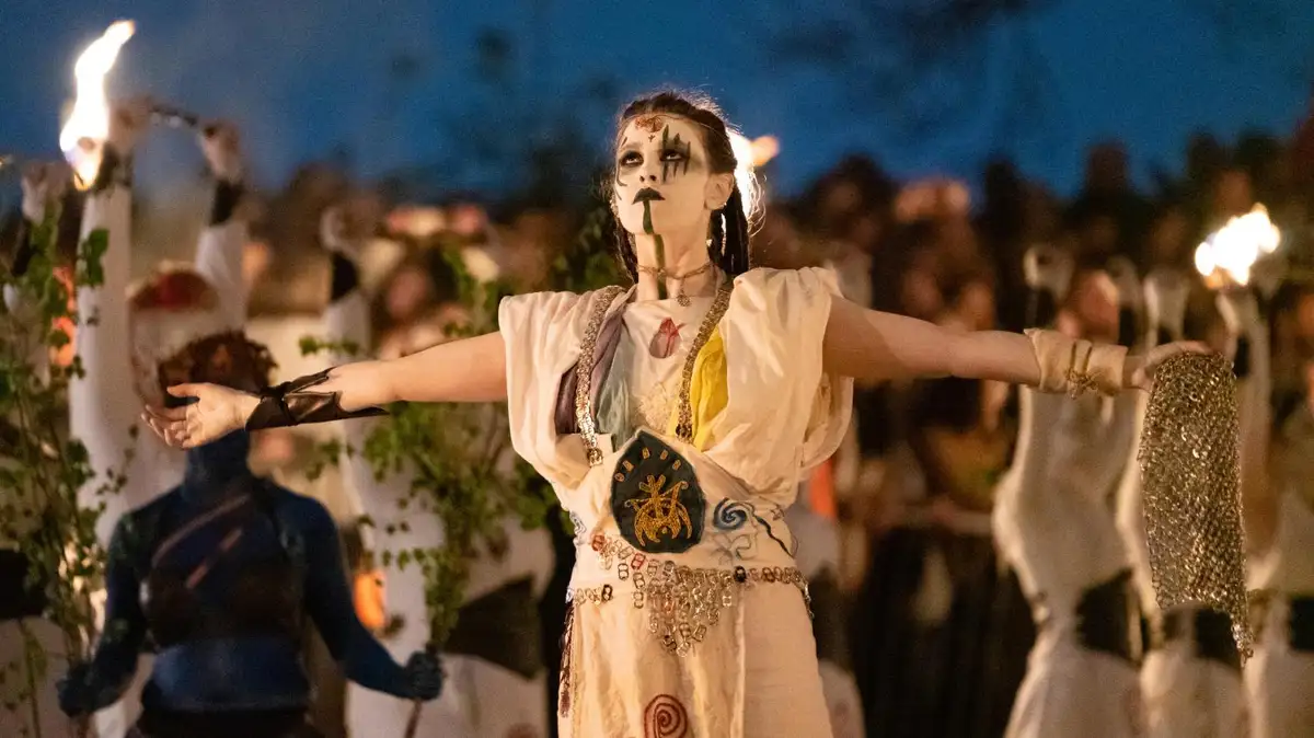 A person in elaborate white ceremonial attire with dramatic black and red face paint, arms outstretched, stands center stage…