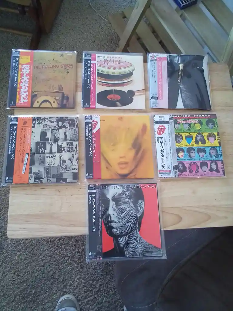 Seven Japanese import CD cases of The Rolling Stones albums are arranged on a wooden table, featuring covers like 'Let It…