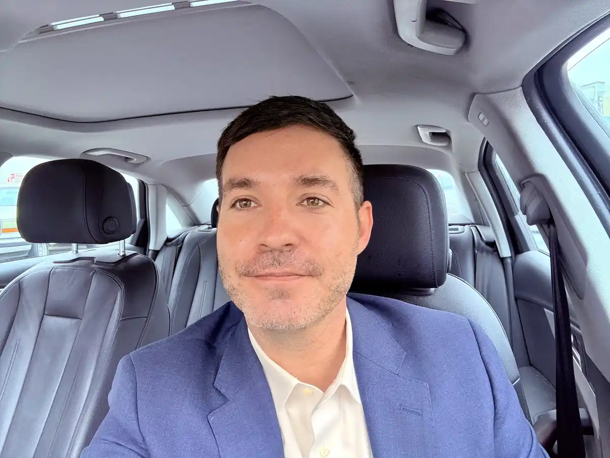A man in a blue suit and white shirt sits in the driver's seat of a car with gray leather interior, looking directly at the…