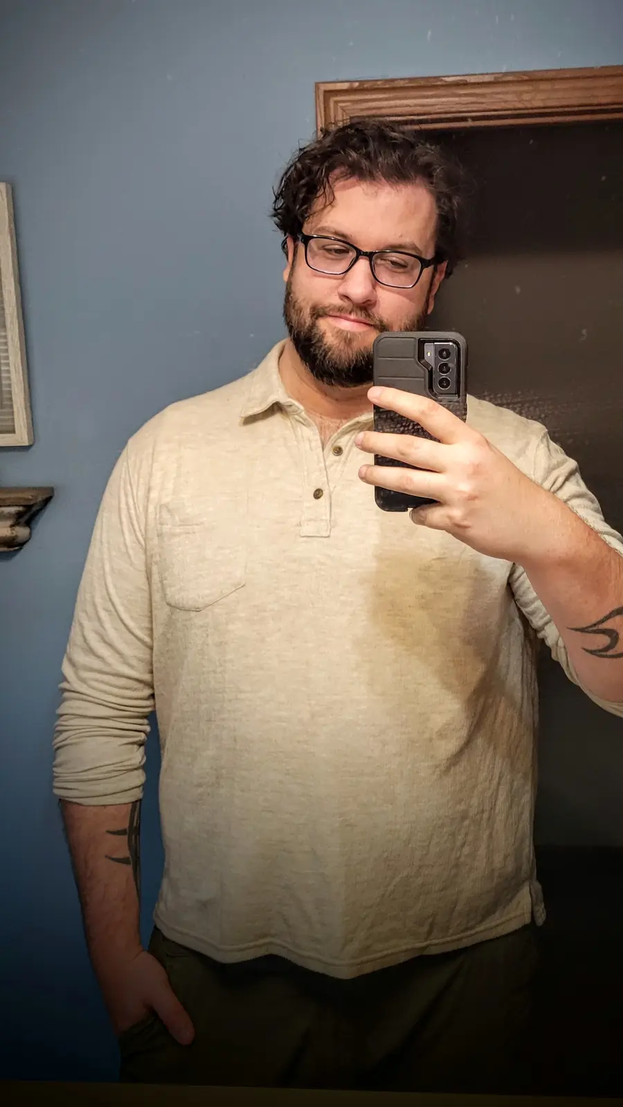 A man with glasses and a beard takes a mirror selfie holding a smartphone, wearing a light-colored long-sleeved shirt that…