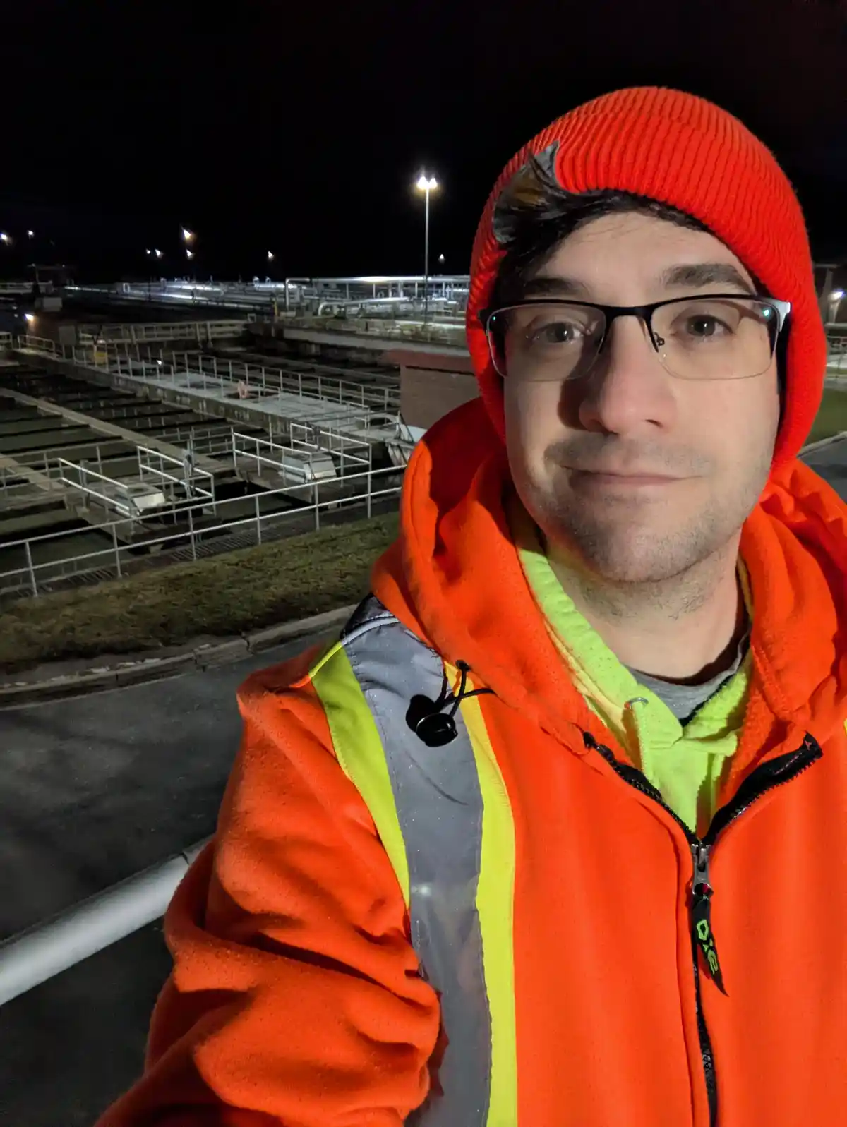 A man wearing an orange beanie and a bright orange high-visibility jacket with reflective stripes takes a selfie at night…