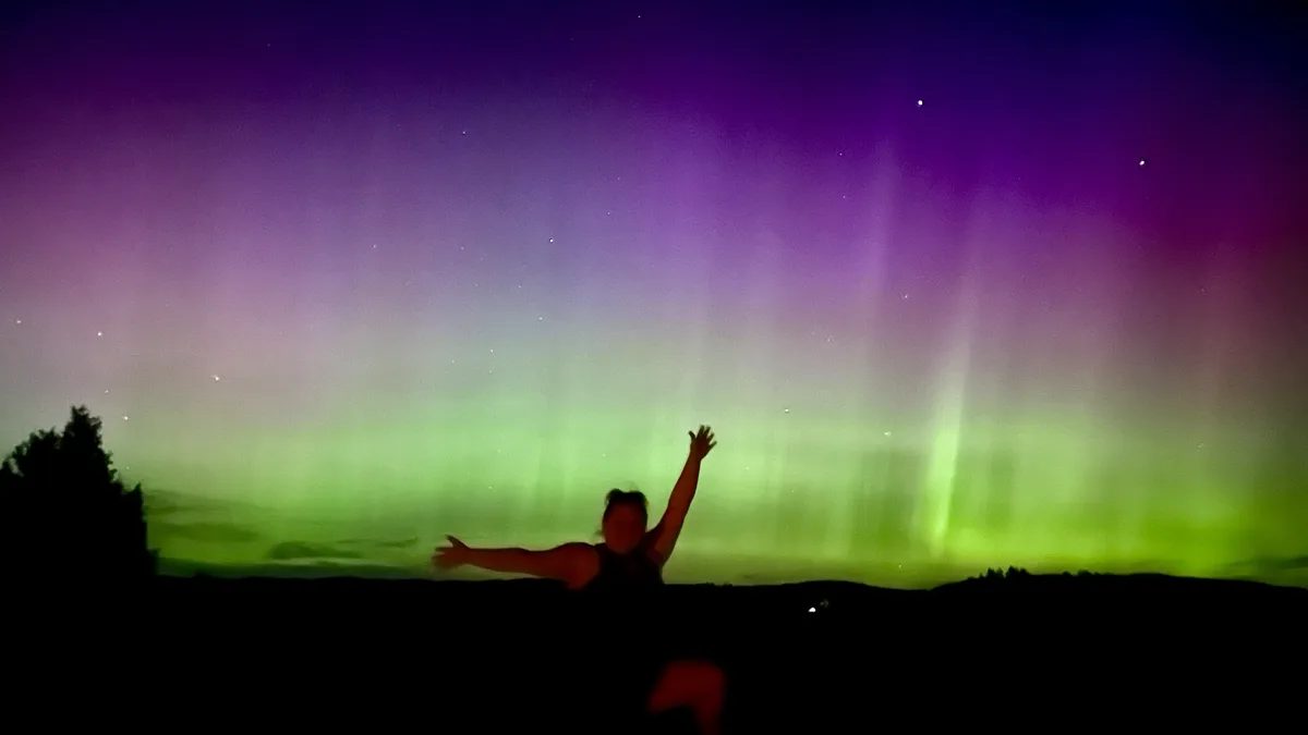 A person is silhouetted against a vibrant aurora borealis, with green and purple lights dancing across the night sky. The…