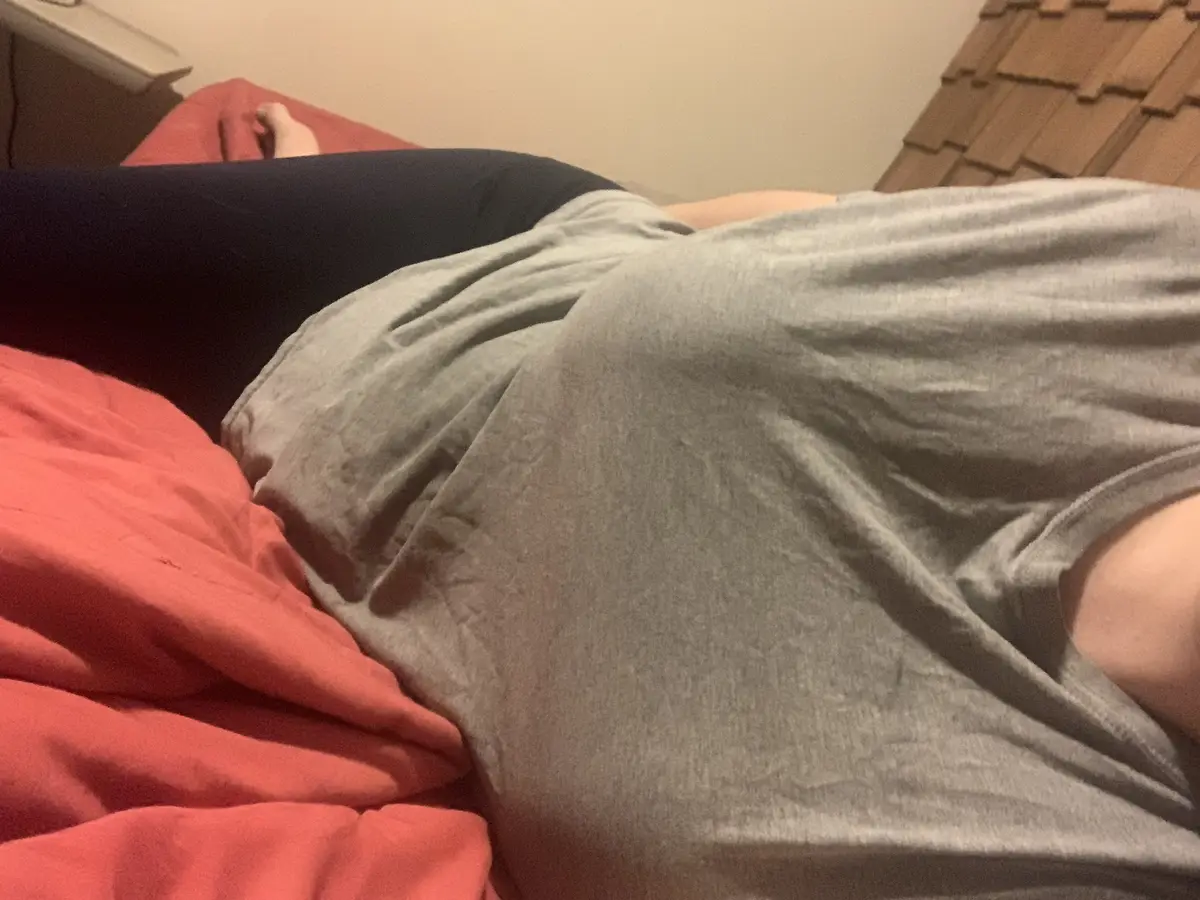 A person is lying on their side on a bed with red sheets, wearing a grey t-shirt and black leggings. The image is a close-up…