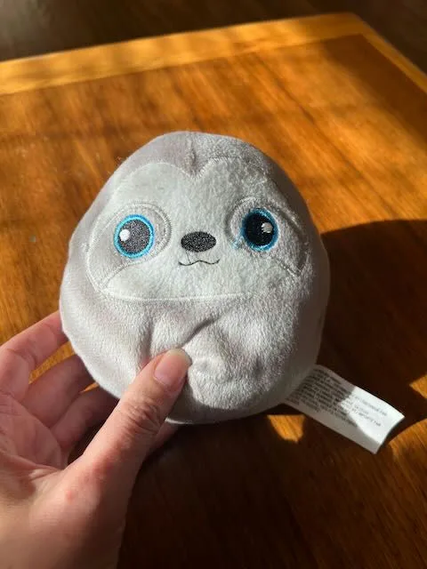 A hand holds a round, gray plush toy with large blue eyes and a small black nose, sitting on a wooden table. The toy…