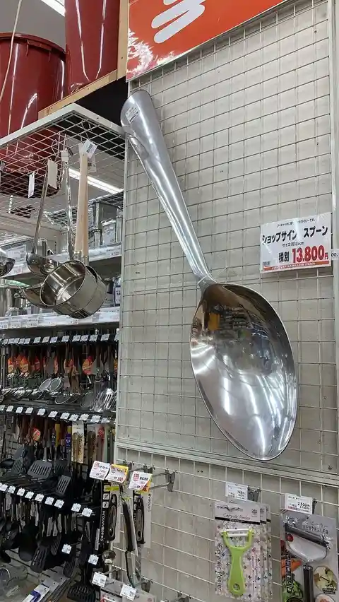 A giant, shiny metal spoon, approximately 125cm long, is mounted on a tiled wall in a Japanese kitchenware store, priced at…