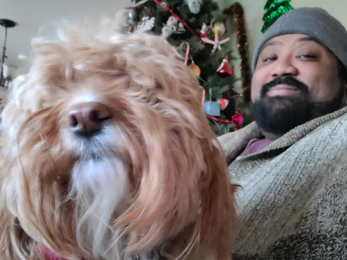 A close-up selfie shows a fluffy, light-brown dog with long hair obscuring its eyes, positioned in front of a man who is…