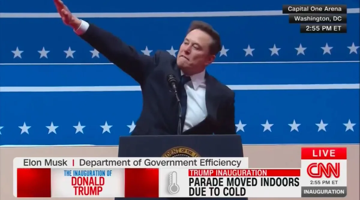 A man in a dark suit and tie stands at a podium with a presidential seal, raising his right arm in a gesture resembling a…