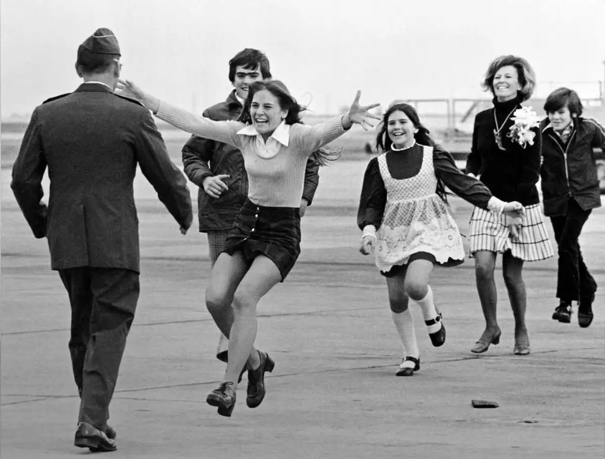 A black-and-white photo shows a group of people, including a woman in a sweater and skirt, joyfully running toward a man in…
