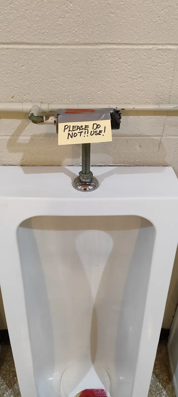 A close-up photo shows a white public urinal with a handwritten note taped to its plumbing. The note, on yellow paper, reads…