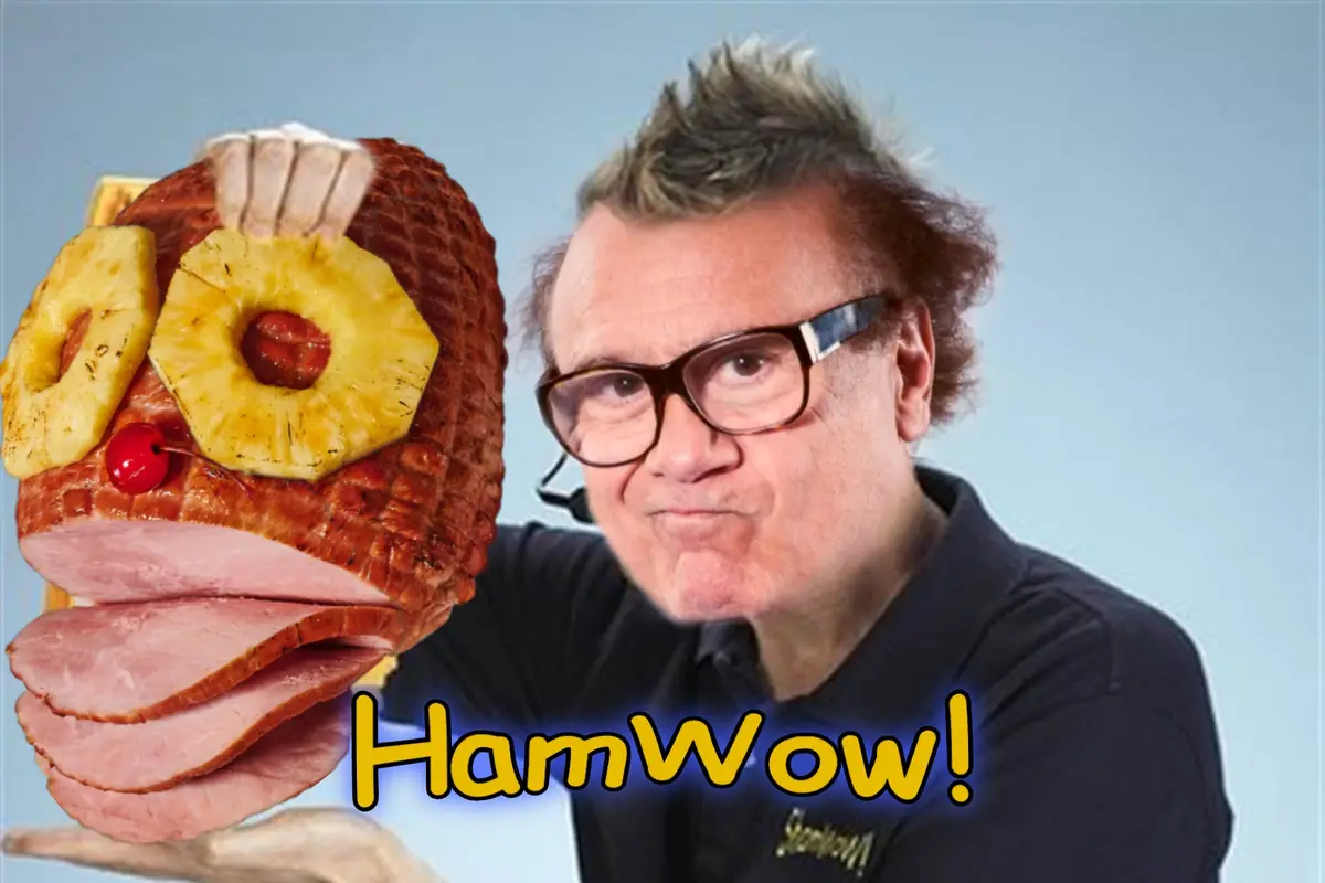 A man with spiky hair and glasses holds up a carved ham decorated with pineapple rings for eyes, a cherry for a nose, and…