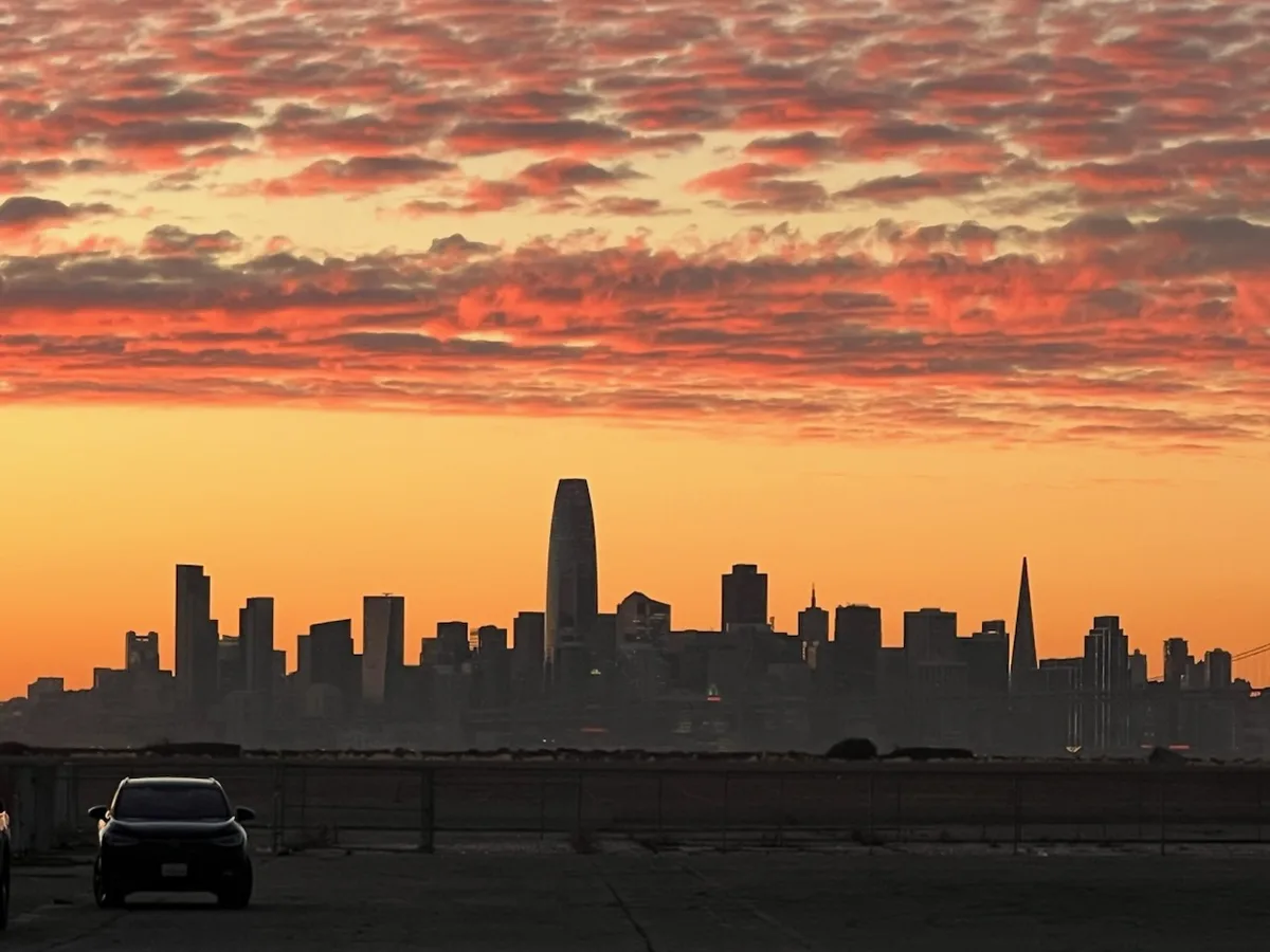 A silhouette of a city skyline at sunset, with vibrant orange and red clouds above. A dark car is parked in the foreground…