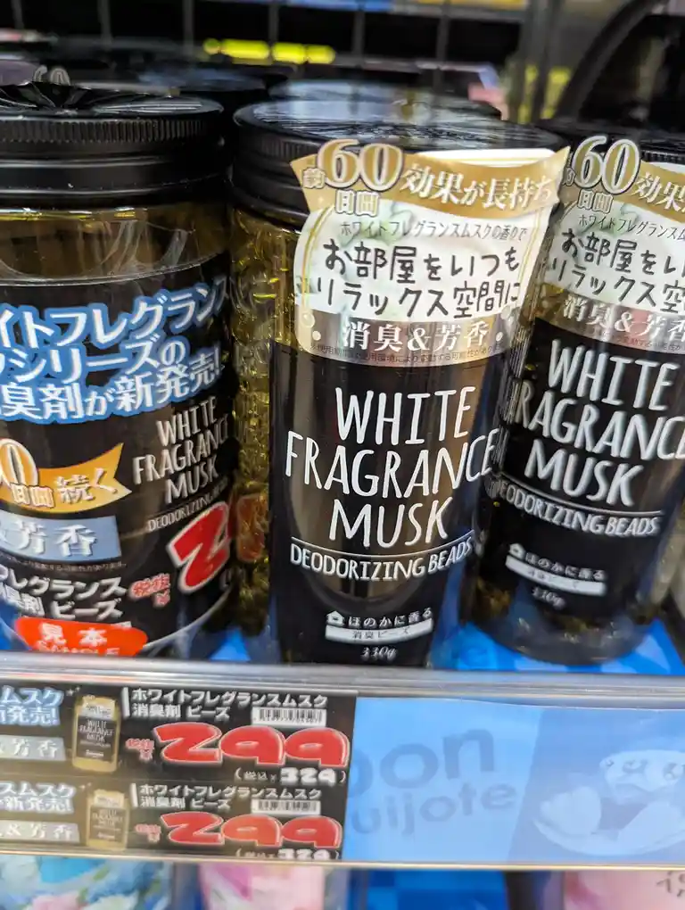 Several black cylindrical containers of 'White Fragrance Musk Deodorizing Beads' are lined up on a store shelf, with…