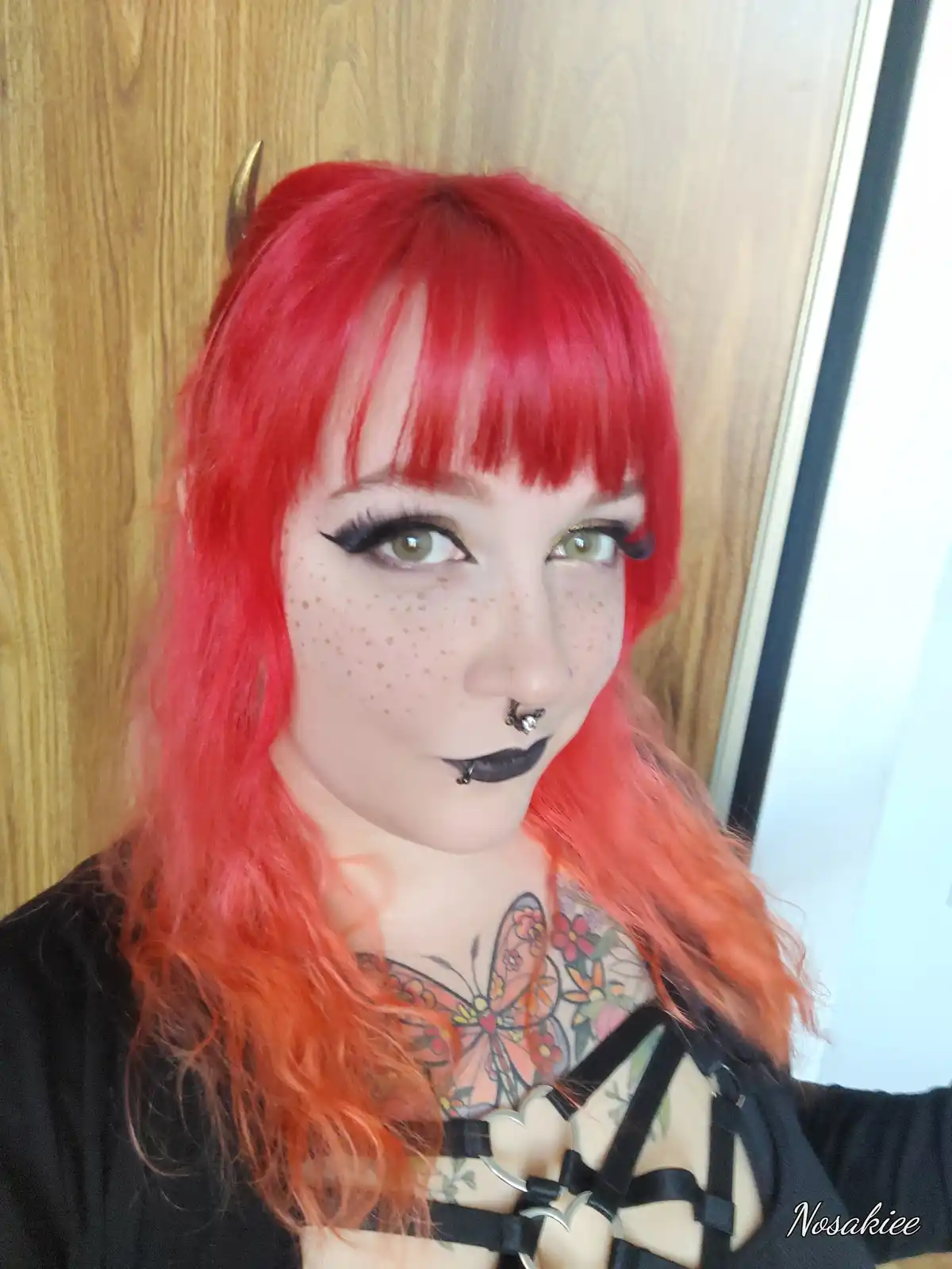 A person with bright red hair and small golden horns poses against a wooden door. They have freckles, black lipstick, a nose…