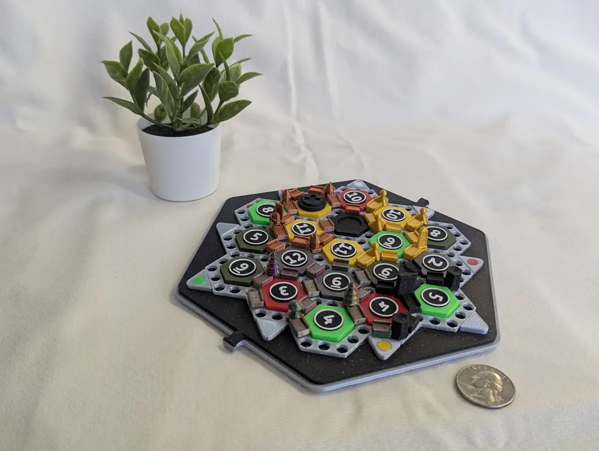 A hexagonal game board with colorful hexagonal tiles numbered 2-12, miniature buildings, and tiny figures is placed on a…