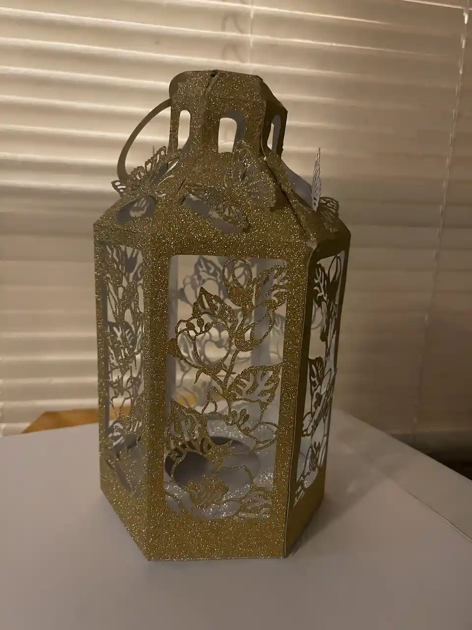 A glittery gold paper lantern with floral cutouts sits on a white surface in front of blinds. The lantern has a handle and…