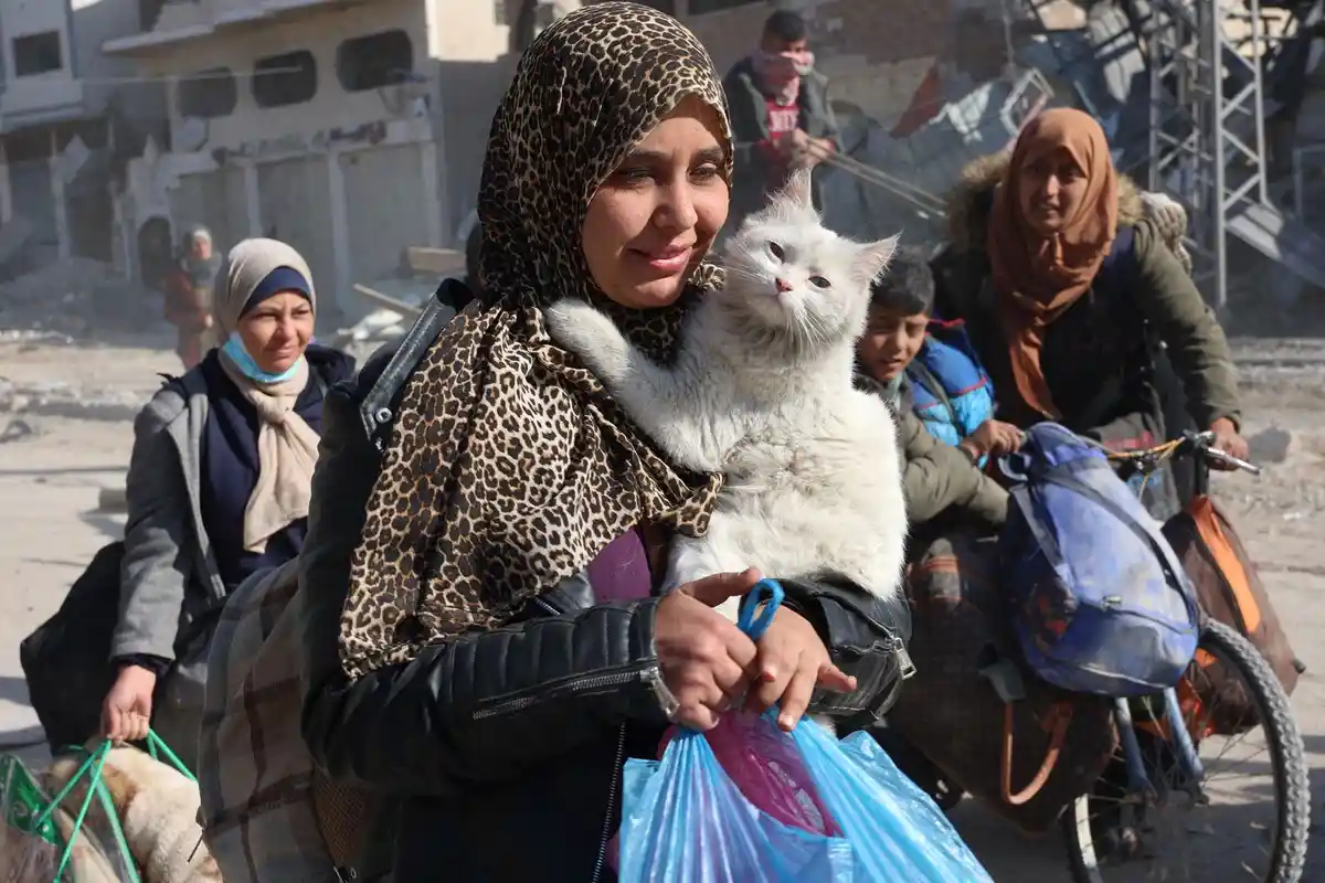 A woman in a leopard-print hijab holds a white cat while standing in a rubble-strewn area with other displaced people and…