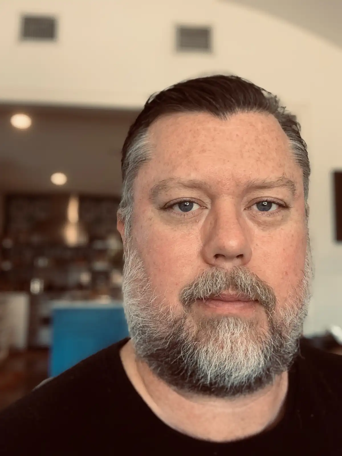 A close-up selfie of a middle-aged man with a graying beard and hair, wearing a black t-shirt, looking directly at the…