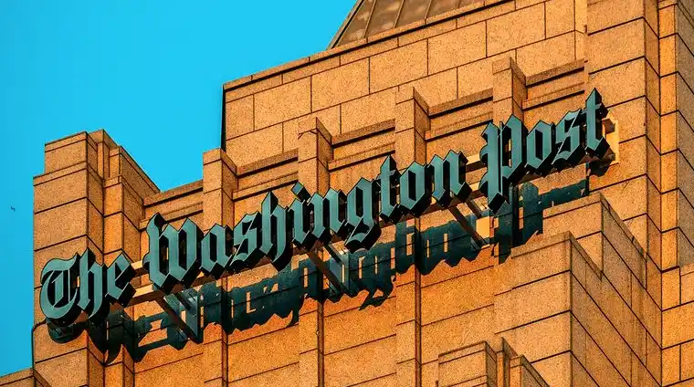 A low-angle photo shows the iconic teal sign of 'The Washington Post' mounted on a tan brick building against a clear blue…