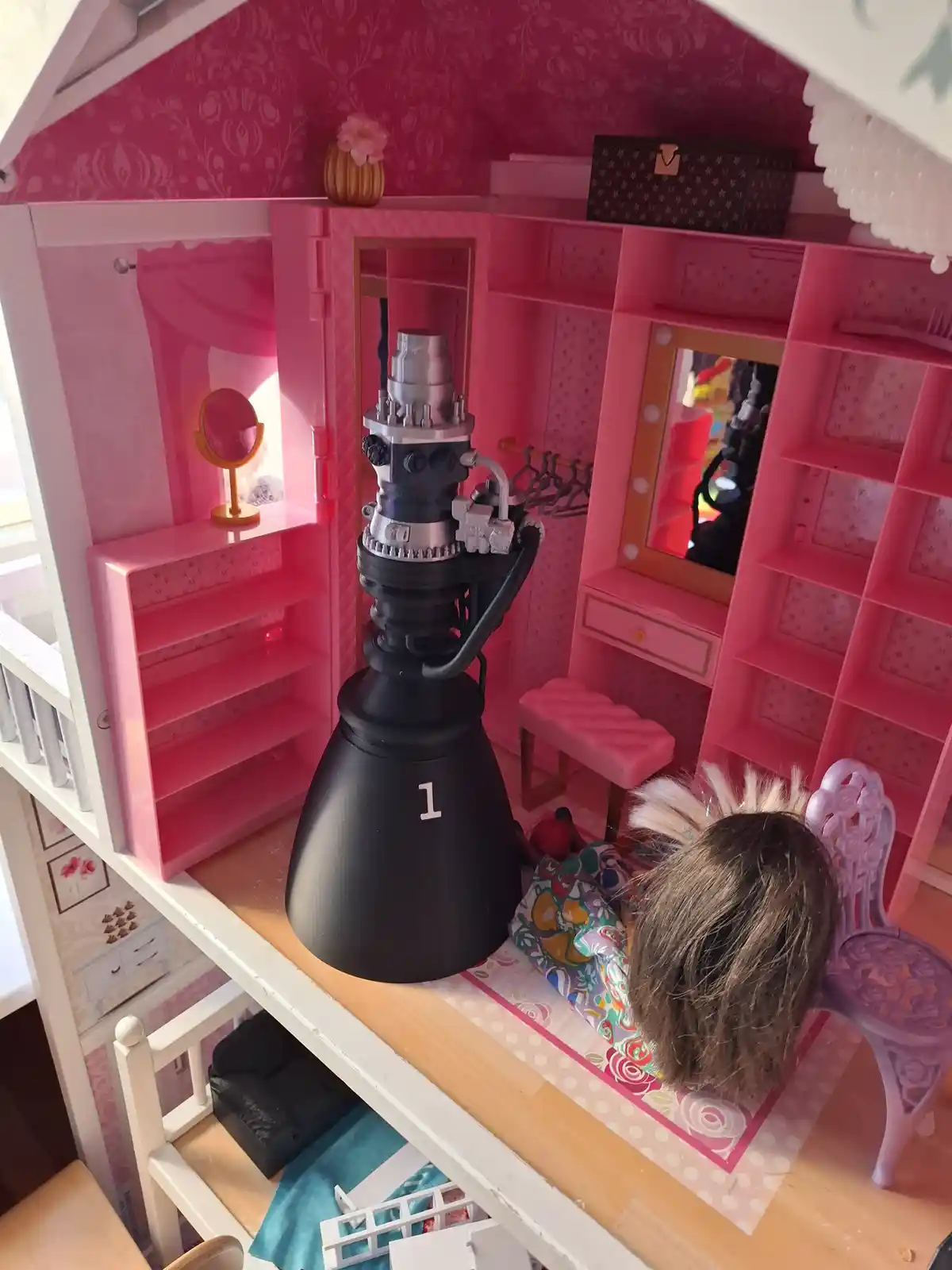 A dollhouse bedroom with pink walls and furniture, featuring a large, realistic-looking rocket engine labeled '1' sitting…