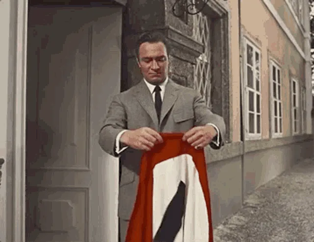 A six-panel sequence shows a man in a grey suit unfolding a red, white, and black flag, which resembles a Nazi flag, from a…