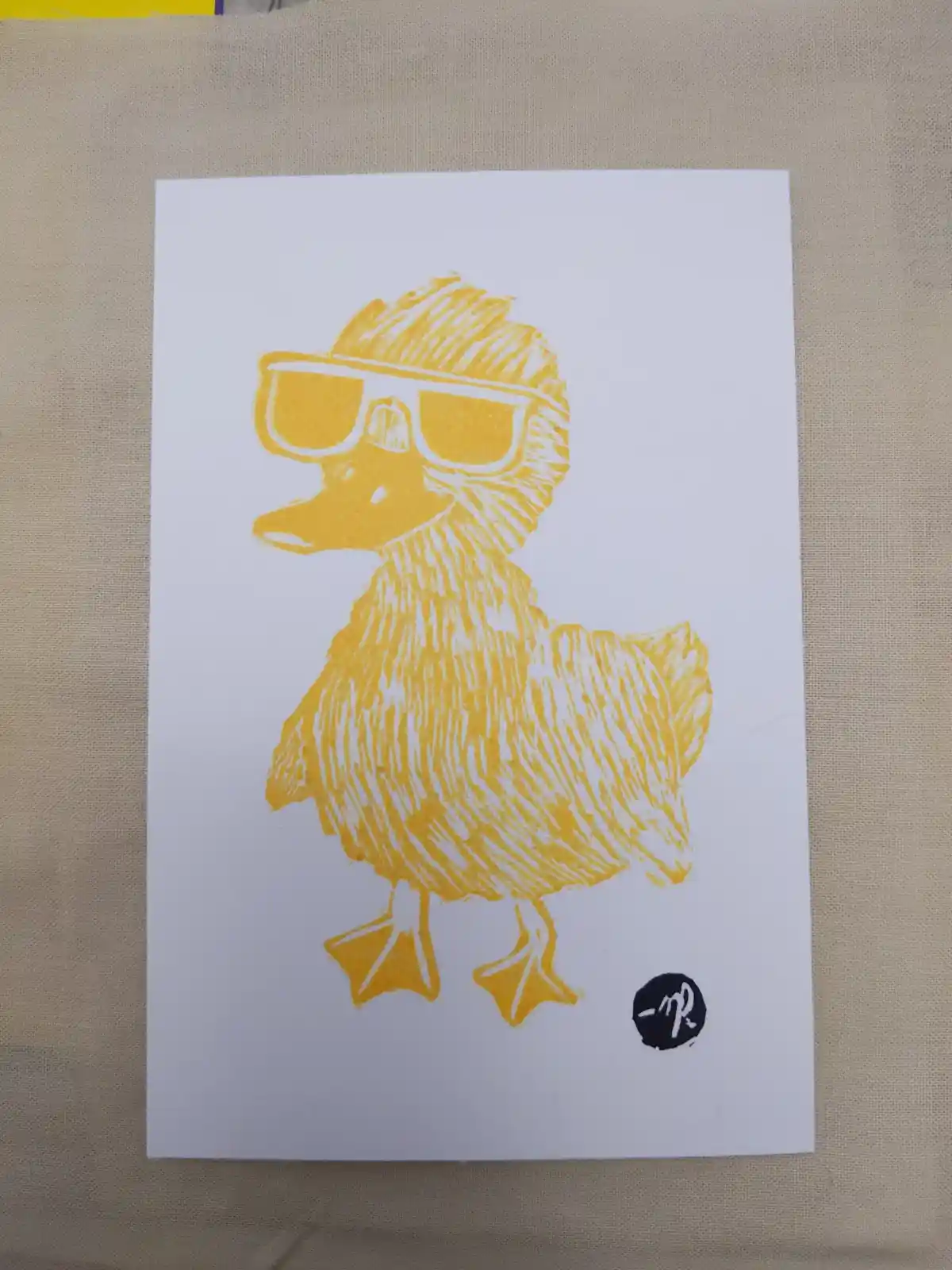 A yellow linocut-style print of a duckling wearing sunglasses and a hat, standing on a white card. The duckling has a…