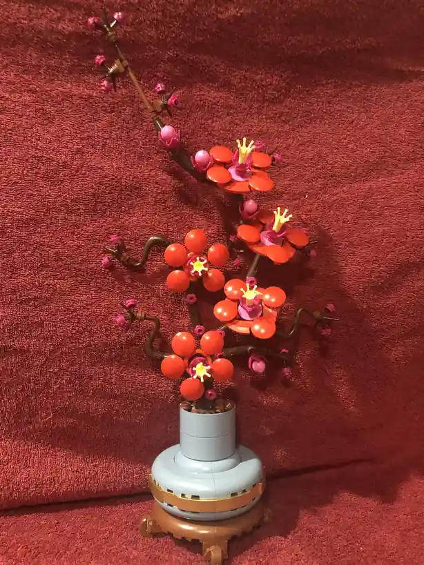 A LEGO model of a cherry blossom tree with red and pink flowers and brown branches, displayed in a light blue and gold base…