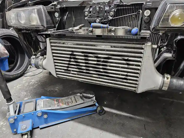 A car's front end is shown with its hood open, revealing a large aftermarket intercooler mounted where the radiator was…