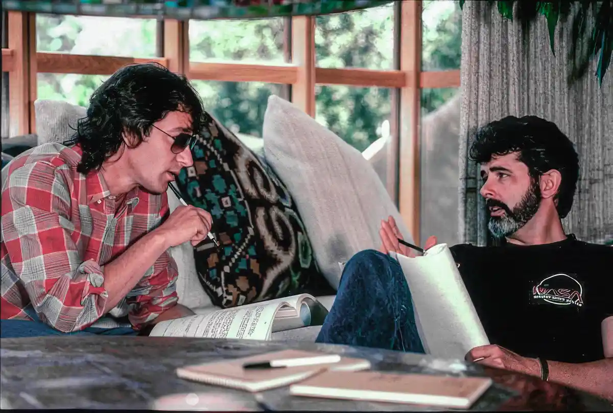 Two men sit on a couch in a room with large windows; one in a red plaid shirt and sunglasses holds a pen to his mouth while…