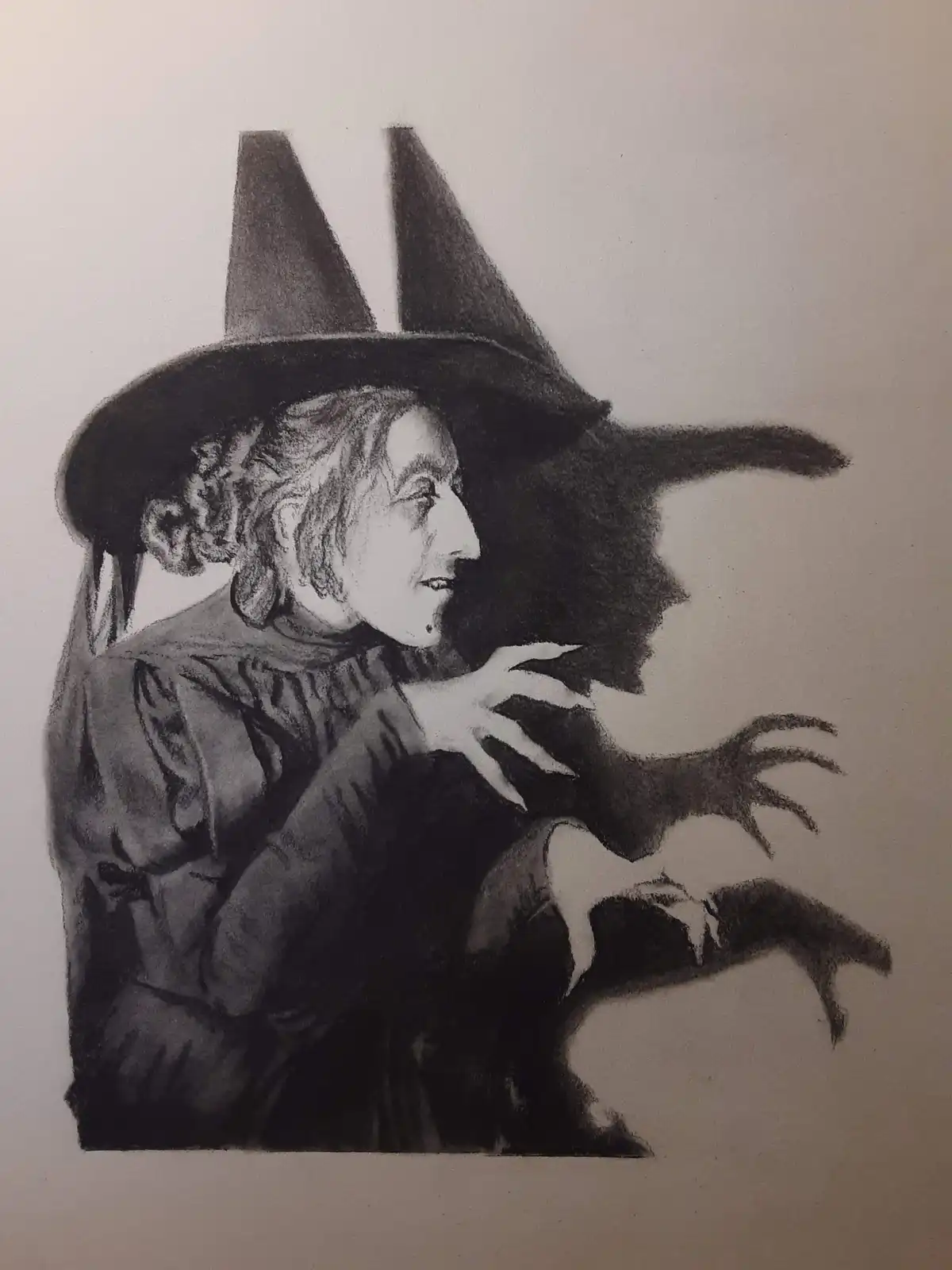 A charcoal drawing depicts a smiling witch in a pointed hat, with her shadow cast behind her, forming the silhouette of a…